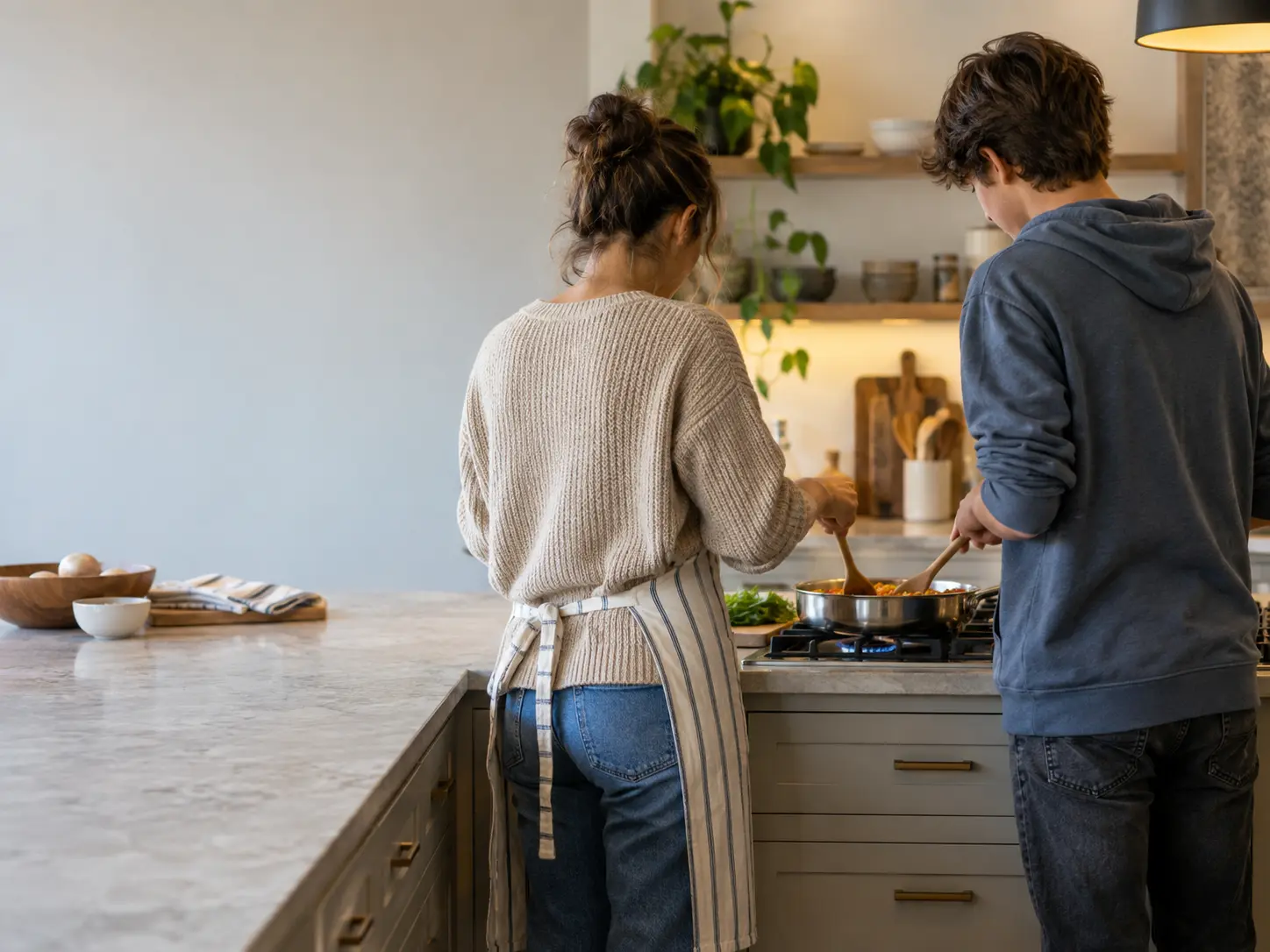 A parent and a teenager stirring a pan together over a gas cooktop set into a kitchen island