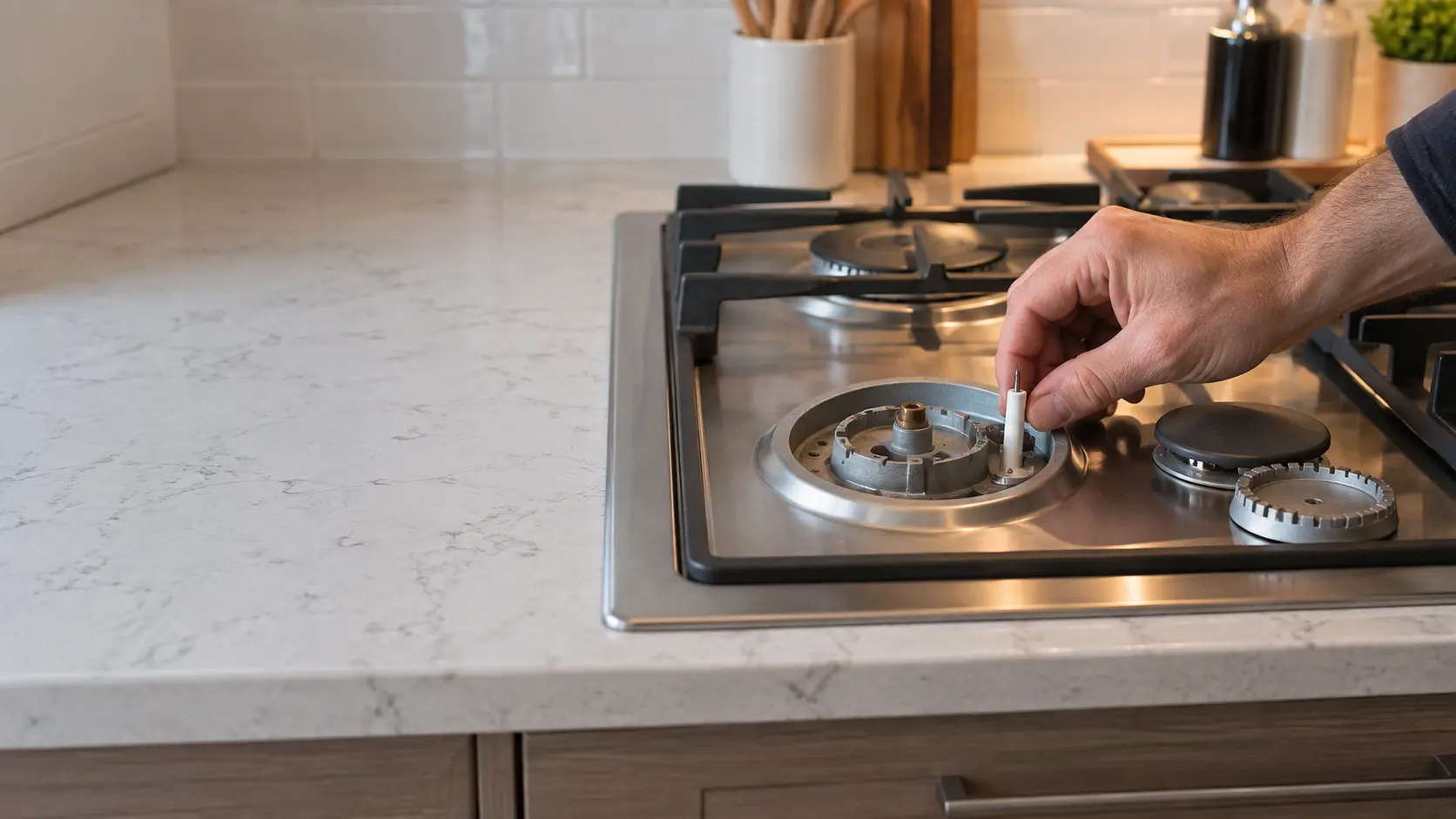 A technician's hand presses a new white ceramic electrode into place beside a bare burner base on a stainless gas cooktop built into a countertop in a US home kitchen.