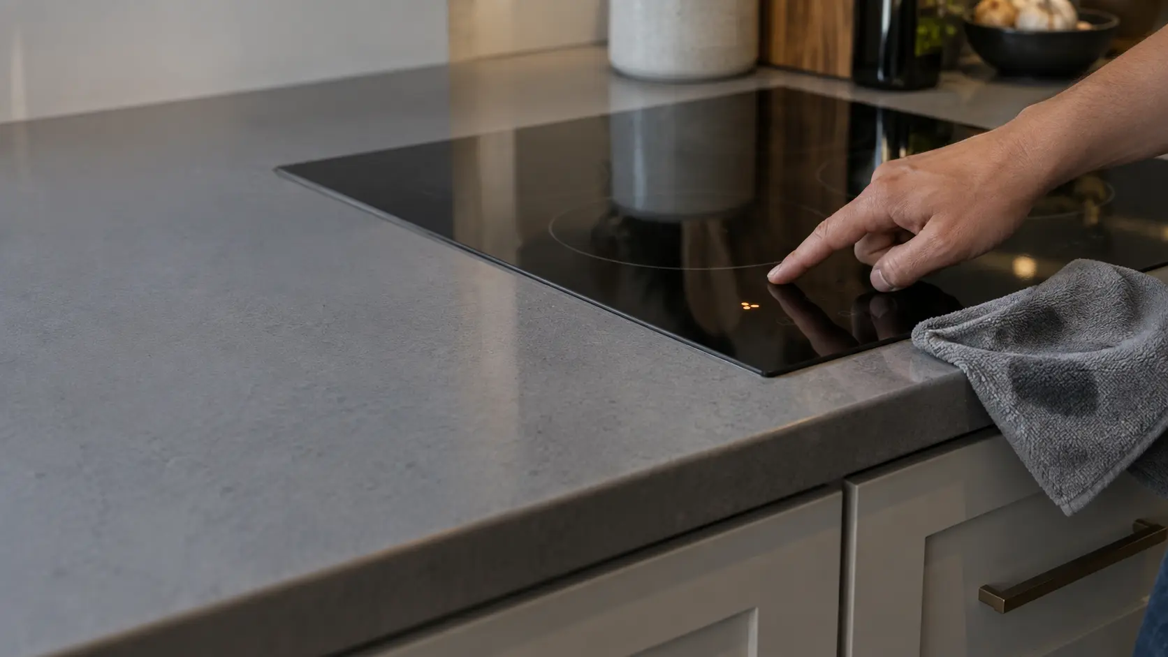 A close view of a finger pressing the touch controls of a black glass cooktop set into a gray quartz countertop, the display dim with one unreadable symbol lit, a damp cloth lying near the keys.
