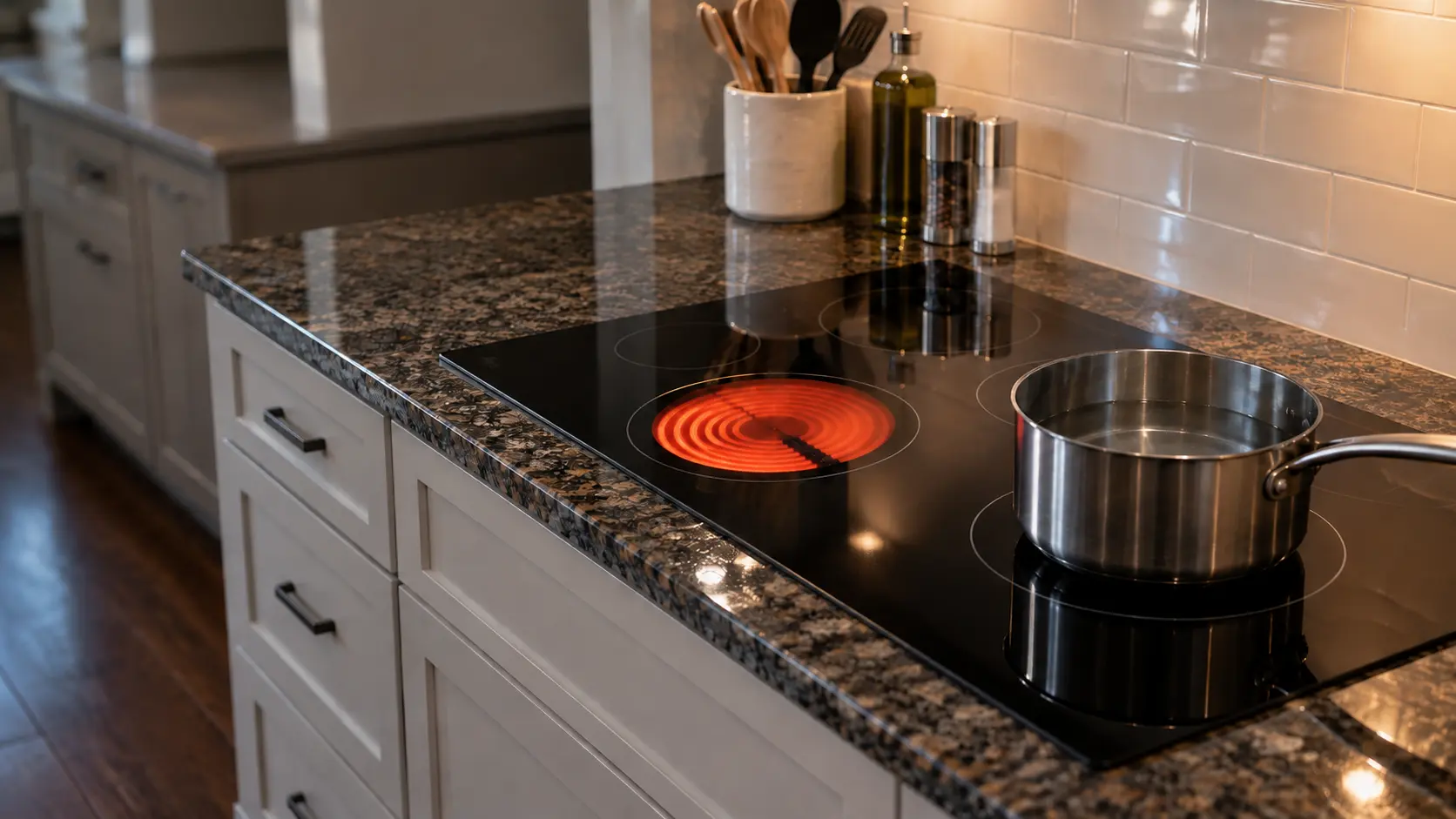 A US home kitchen with a black radiant glass cooktop set into a granite countertop, one ring glowing orange while the ring beside it stays dark under a pot of still water.