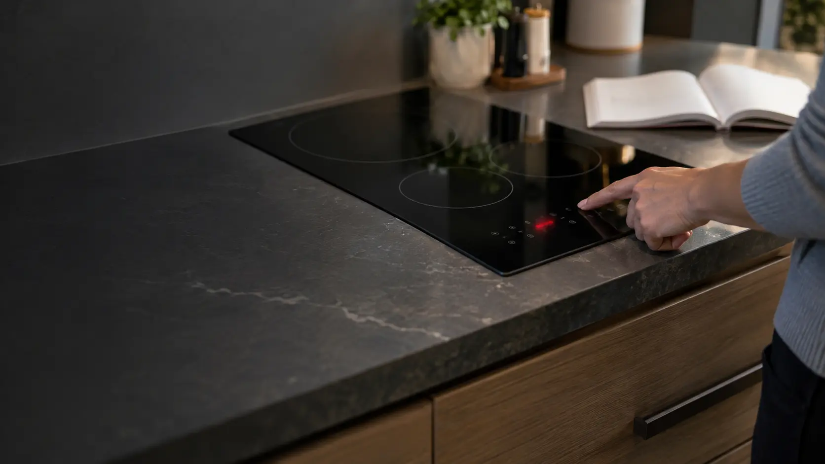 A close view of the touch display on a black induction cooktop set into a stone countertop, a blurred unreadable code glowing on it and an owner's manual lying open beside it with out-of-focus pages.