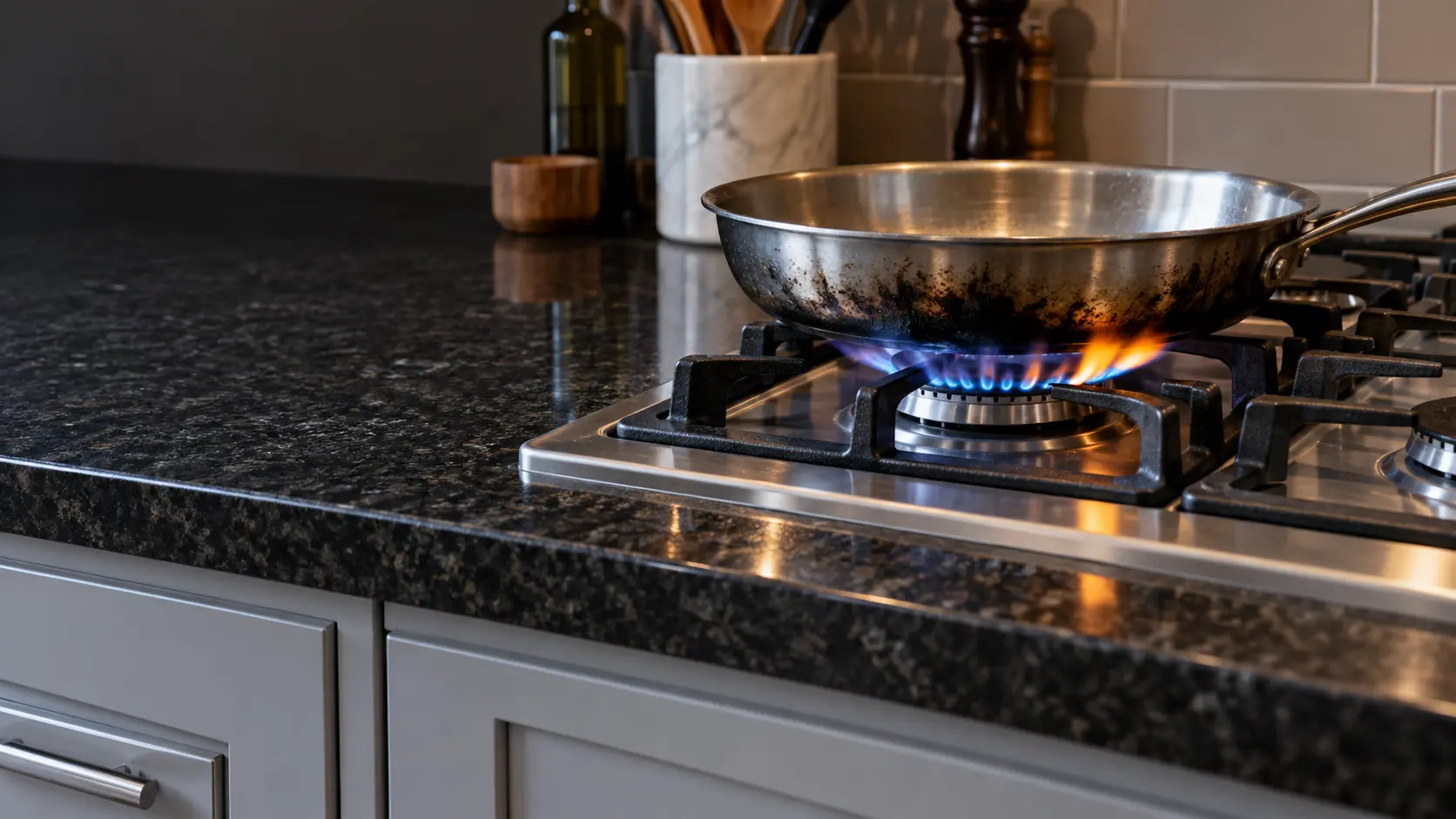 A close view of a lit gas burner on a stainless cooktop set into a dark granite countertop, the flame lazy and yellow-tipped on one side beneath an empty steel pan with soot on its base.