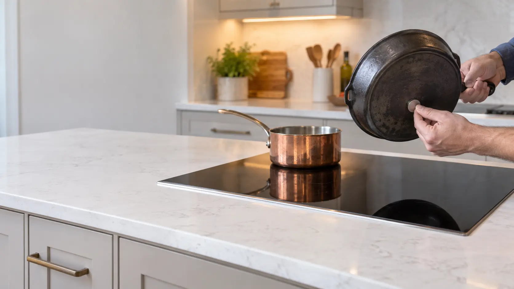 A black induction cooktop set flush into a white quartz countertop in a US home kitchen, a small copper saucepan sitting on a large zone and a hand holding a small magnet to the base of a cast-iron pan beside it.