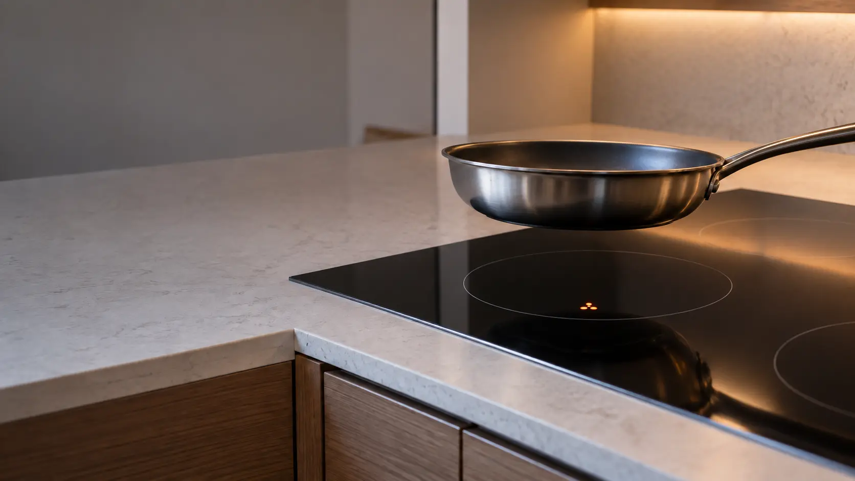 A pan lifted from a cool induction cooktop in a quartz counter, the zone underneath showing a small lit warning letter where the pan had been.