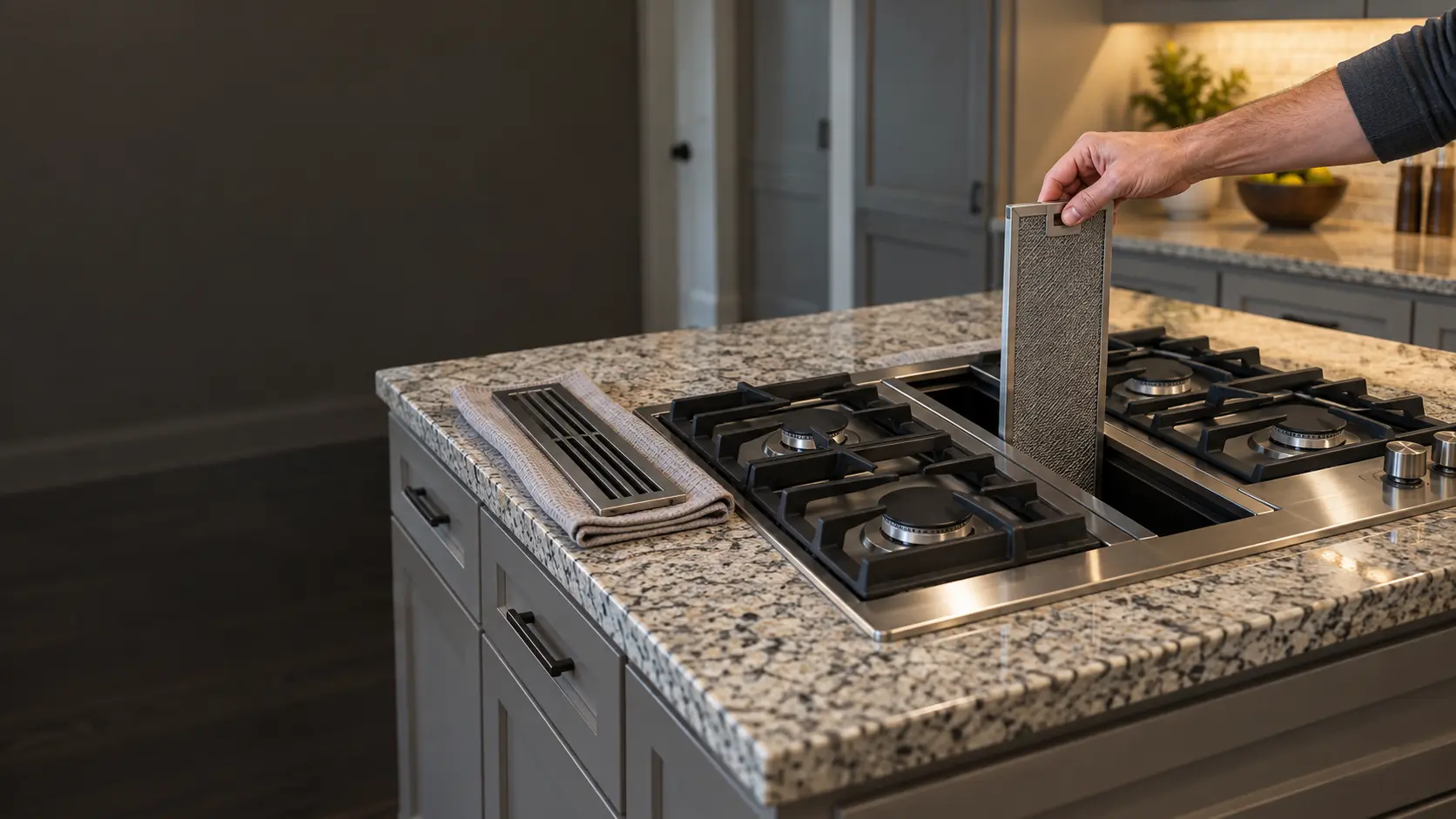 A hand lifting a stainless mesh grease filter out of the center vent strip of a gas downdraft cooktop set into a speckled granite island in a US home kitchen, the vent grate resting on a towel beside it and the burners off.