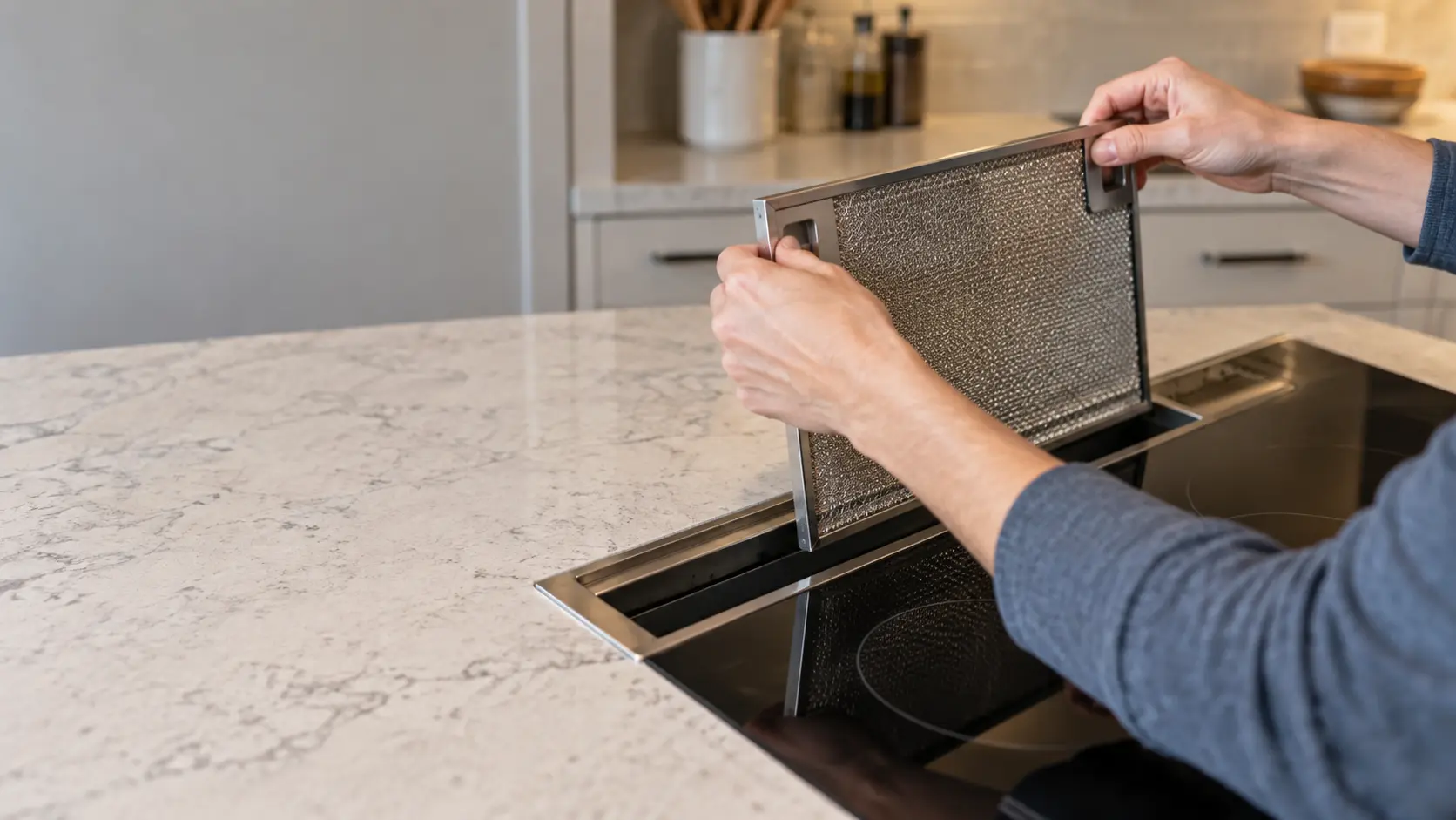 Close on a metal grease filter lifted out of a cooktop's downdraft vent slot, the empty slot below.