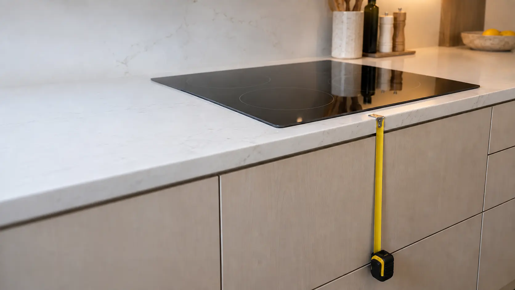 A black induction cooktop set into a quartz counter in a US home kitchen, a tape measure hooked on the counter edge and running straight down the closed flat-panel cabinet face below it.