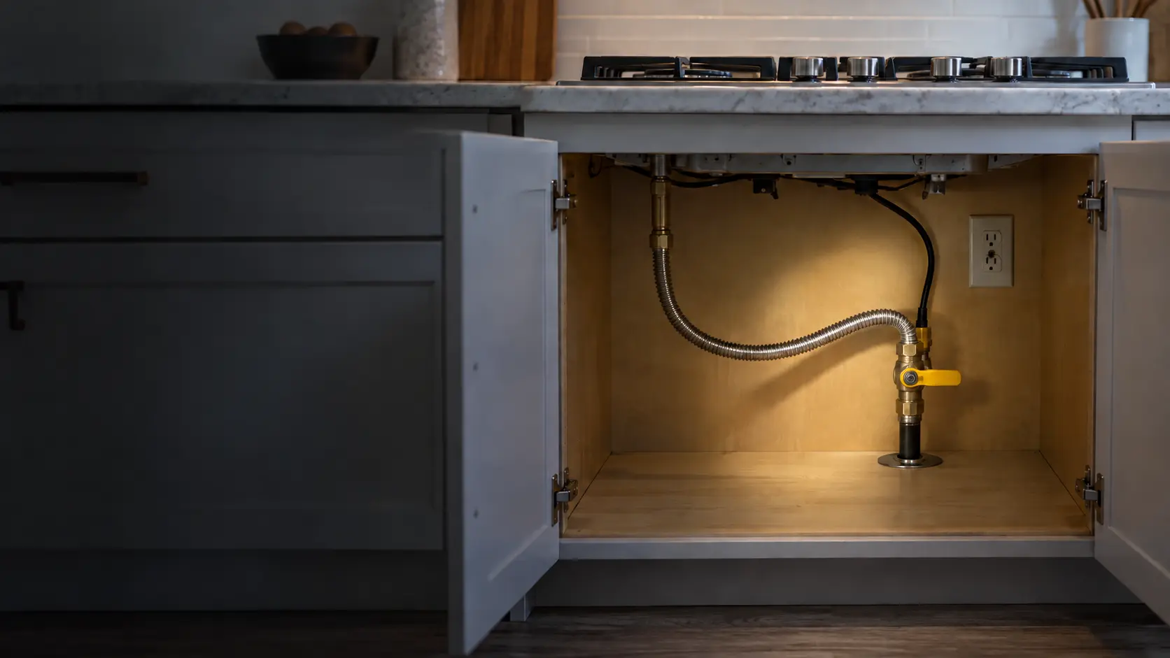 The open base cabinet under a built-in gas cooktop in a US home kitchen, lit by a flashlight, showing a yellow-handled gas shutoff valve, a coiled flexible connector and a small outlet on the cabinet back, with the cooktop's knobs just visible above the counter edge.
