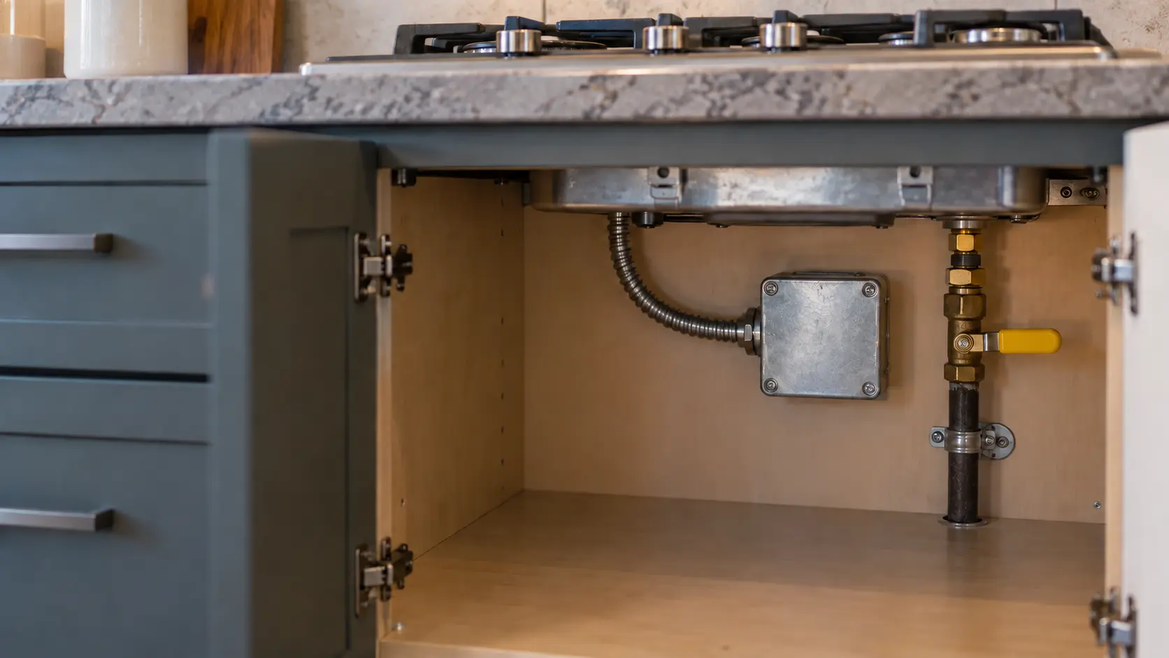 Close on a metal junction box and a gas shutoff valve on the back wall of the cabinet under a cooktop.