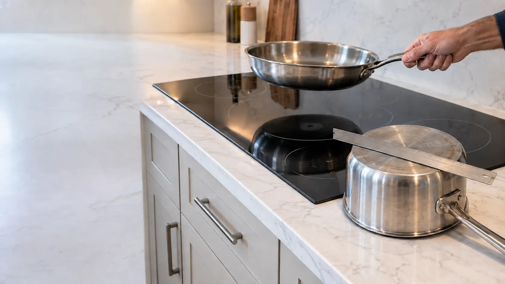 A stainless clad skillet lifted a few inches above a black radiant glass cooktop set into a white marble counter in a US home kitchen, a straight steel ruler lying across the base of an upturned saucepan beside it.