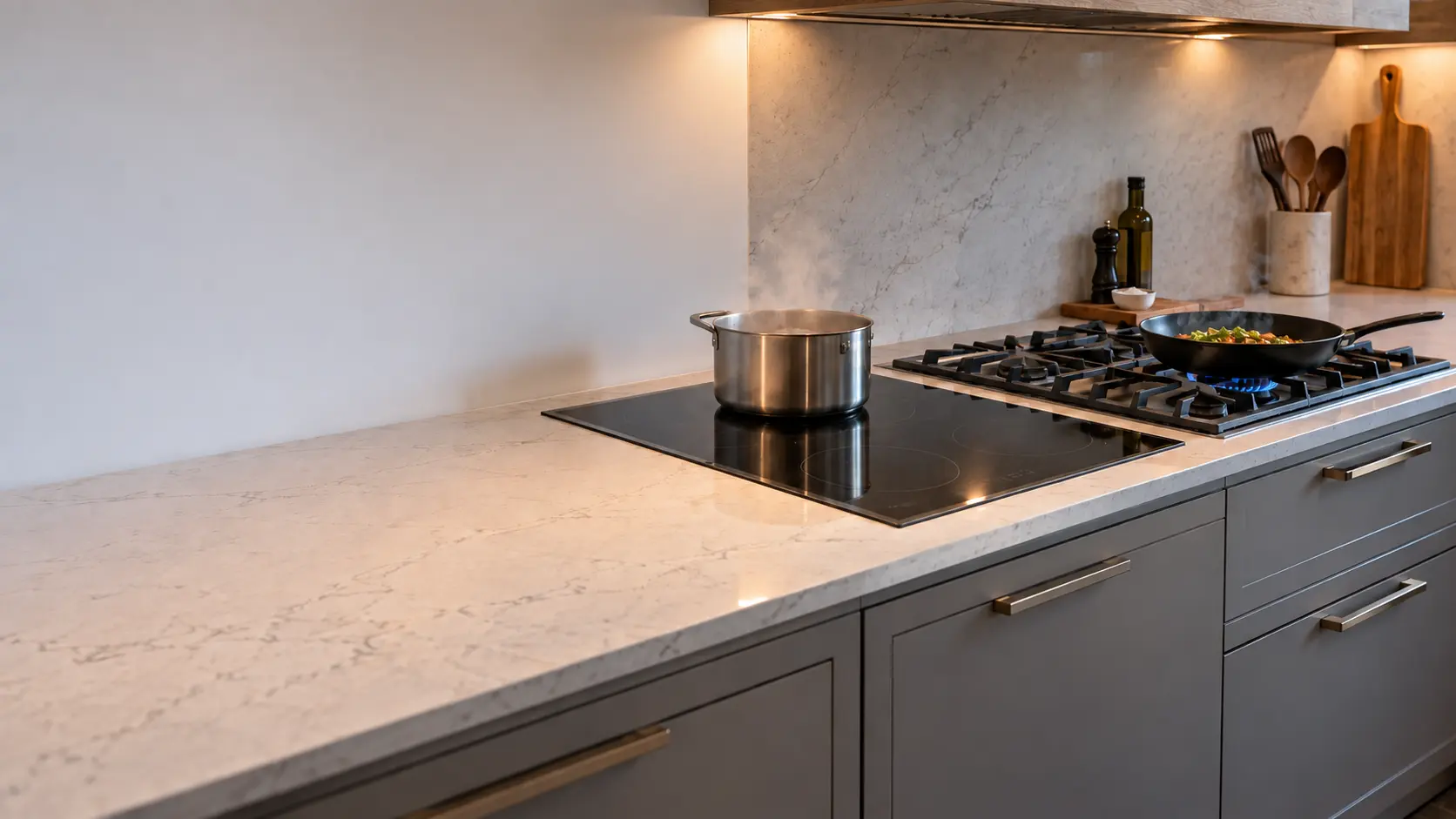 A gas cooktop with cast-iron grates and a flush black induction cooktop set side by side into a long quartz counter in a US home kitchen, drawers below both, one blue flame lit under a skillet and a pot on the induction zone.