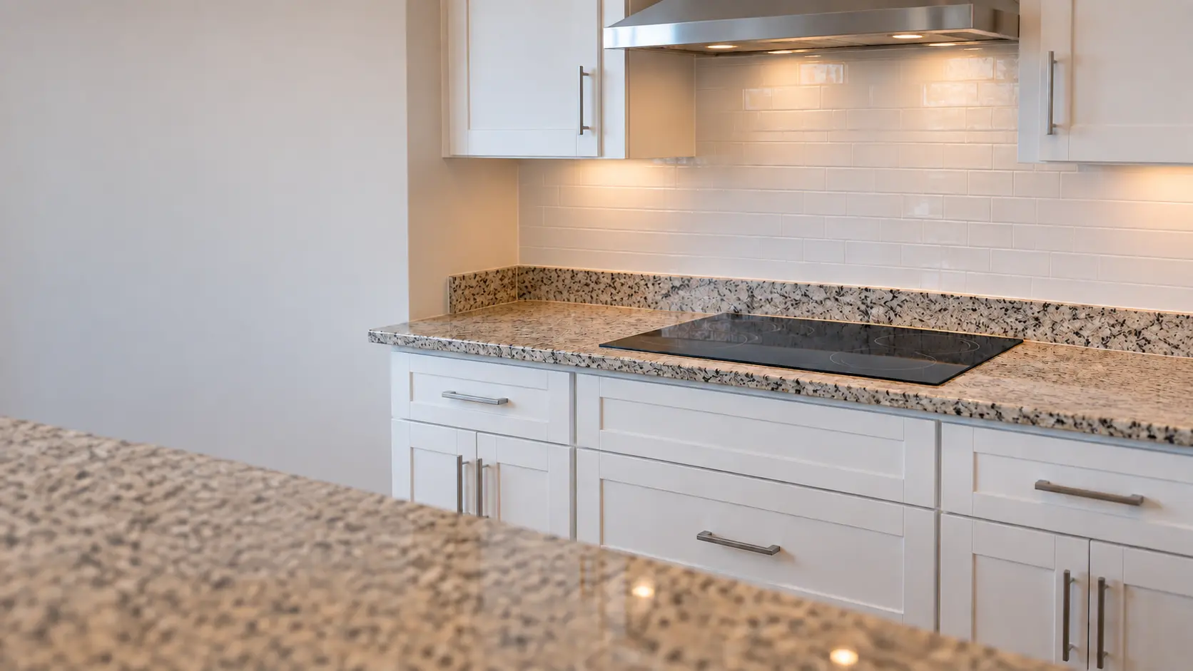 A built-in cooktop in a Florida block house kitchen with a 30-inch glass radiant cooktop in a laminate-edged granite counter and white shaker drawers below, seen from across the room.